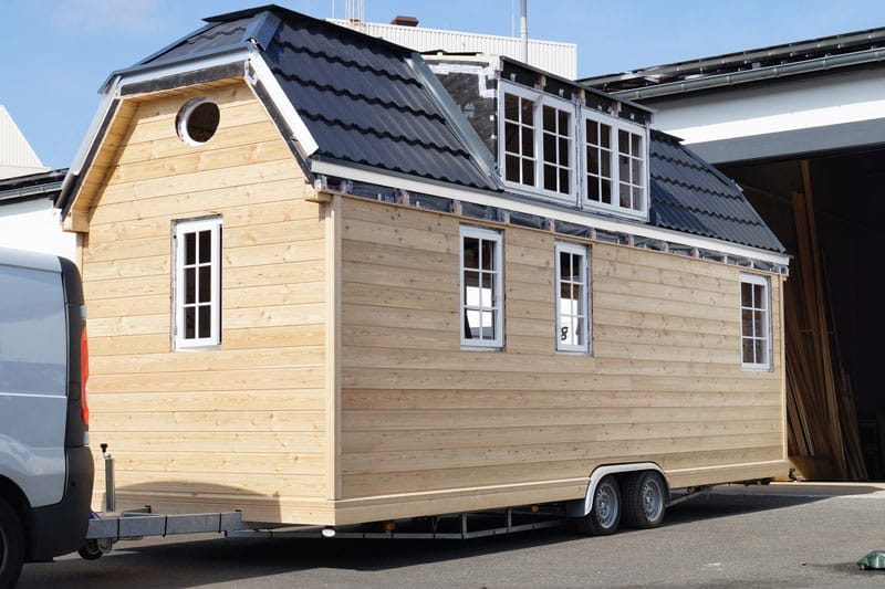 Ein Tiny House mit hellem Holz und dunklem Krüppelwalmdach auf einem Anhänger