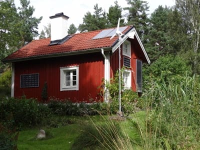 Rotes Häuschen umgeben von Grün mit schwarzen Solarthermie-Kollektoren und Windrad am Giebel