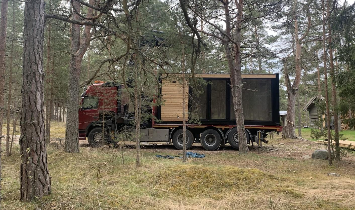Ein LKW liefert ein PopUpHut-Wohnmodul mit Holzverkleidung und großen Fenstern in einem Waldgebiet