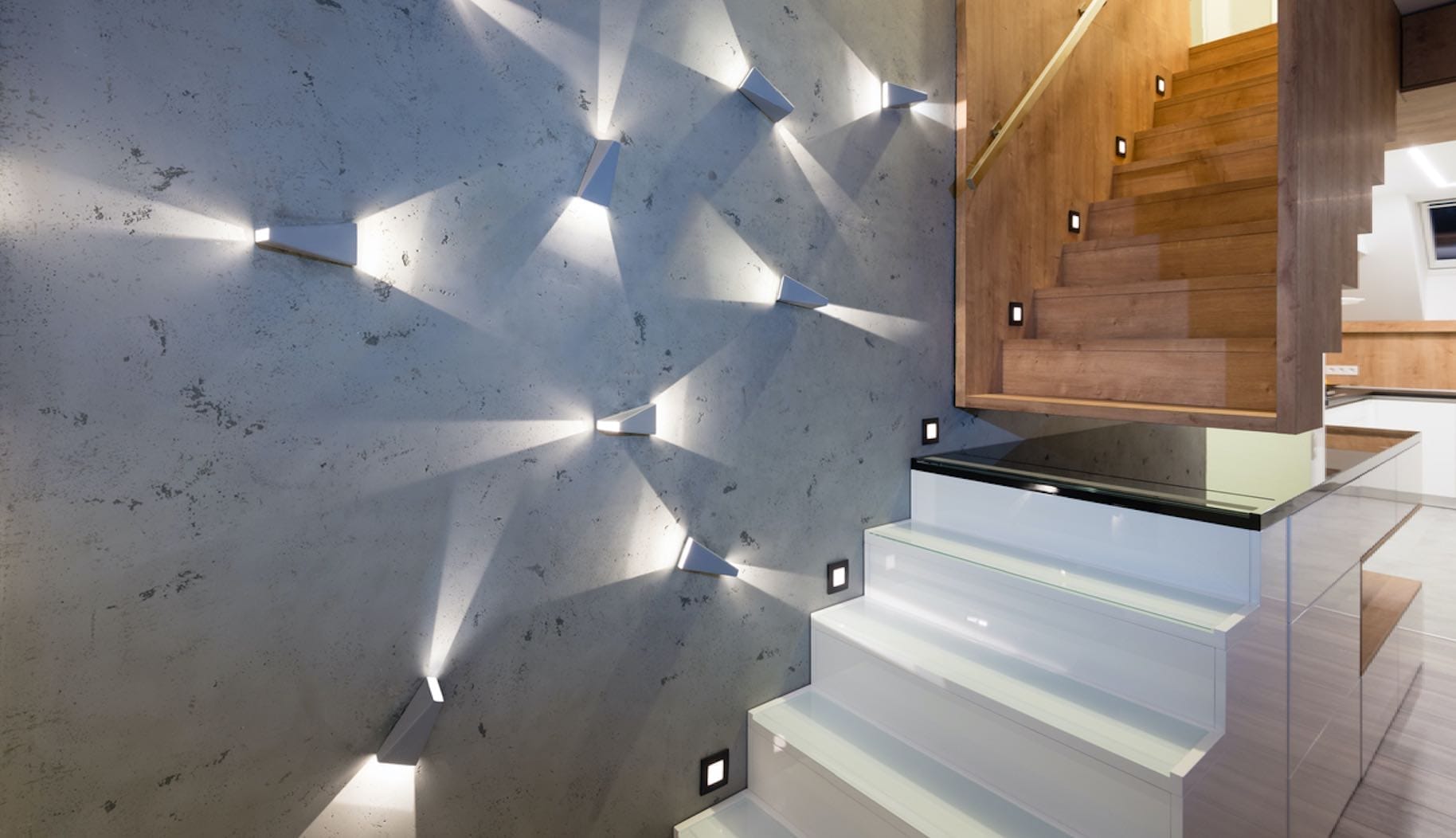 Wandleuchten erzeugen Lichtkegel an einer grauen Wandfläche neben einer modernen Treppe aus Holz und Glas