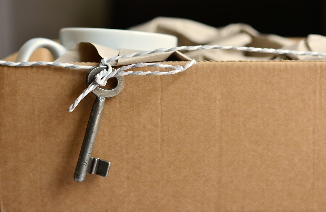 Ein alter Schlüssel hängt an einer Schnur über einem offenen Umzugskarton mit verpacktem Geschirr