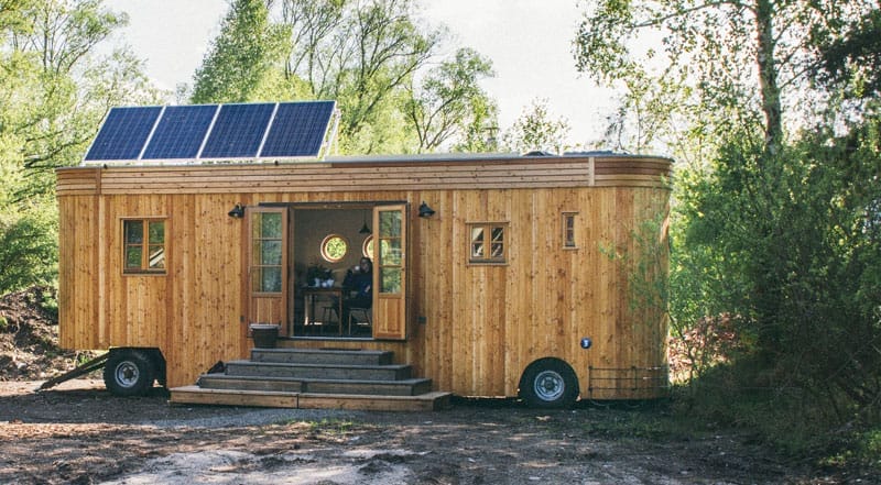 Holziges Tiny House auf Rädern mit Solarpaneelen auf dem Dach, umgeben von grünen Bäumen