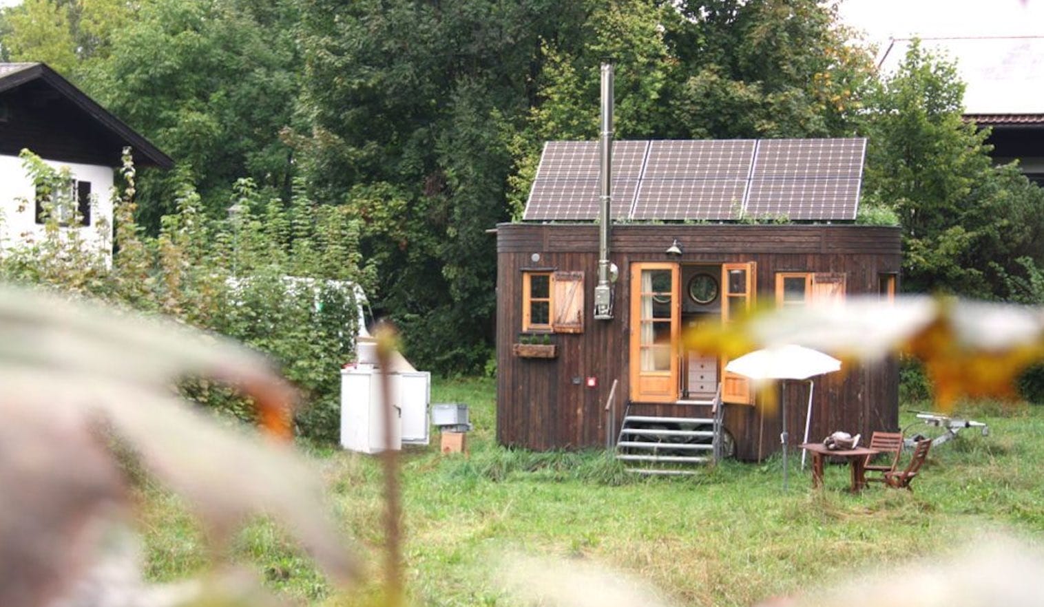 Holz-Tiny-House mit Solarmodulen auf dem Dach und einem hohen Ofenrohr, daneben kleiner Tisch mit 2 Stühlen