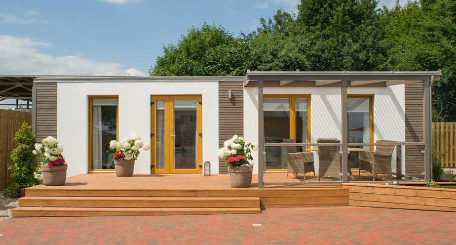 Modernes Modulhaus mit großer Holzterrasse, Glasgeländer und beiger Terrassenüberdachung