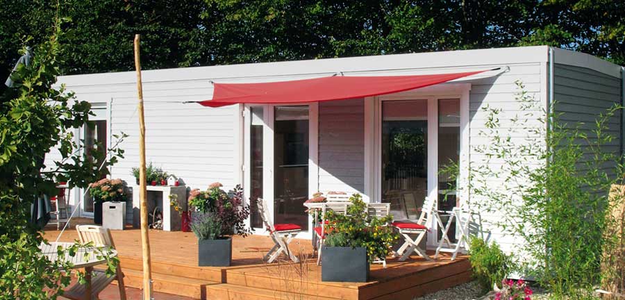 Hausfassade mit großer Holzterrasse, auf der Gartenmöbel stehen und ein rotes Sonnensegel gespannt ist