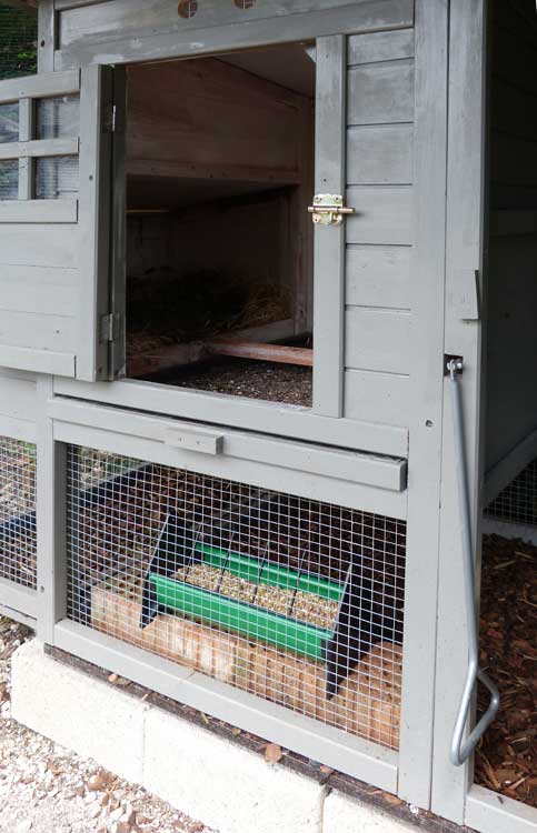 Nahaufnahme eines grauen Hühnerstalls mit geöffneter Tür, im Inneren mit Heu und Einstreu, darunter ein Futtertrog