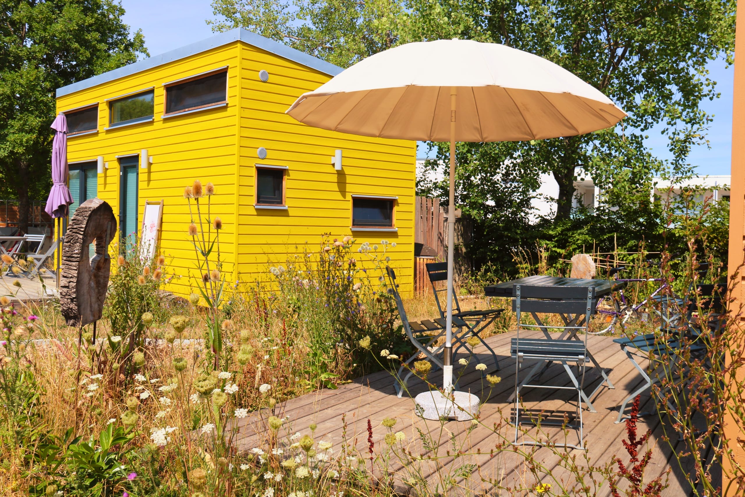 Gelbes Tiny House mit Terrasse, Sitzgelegenheiten und einem Sonnenschirm in einer naturnahen Umgebung