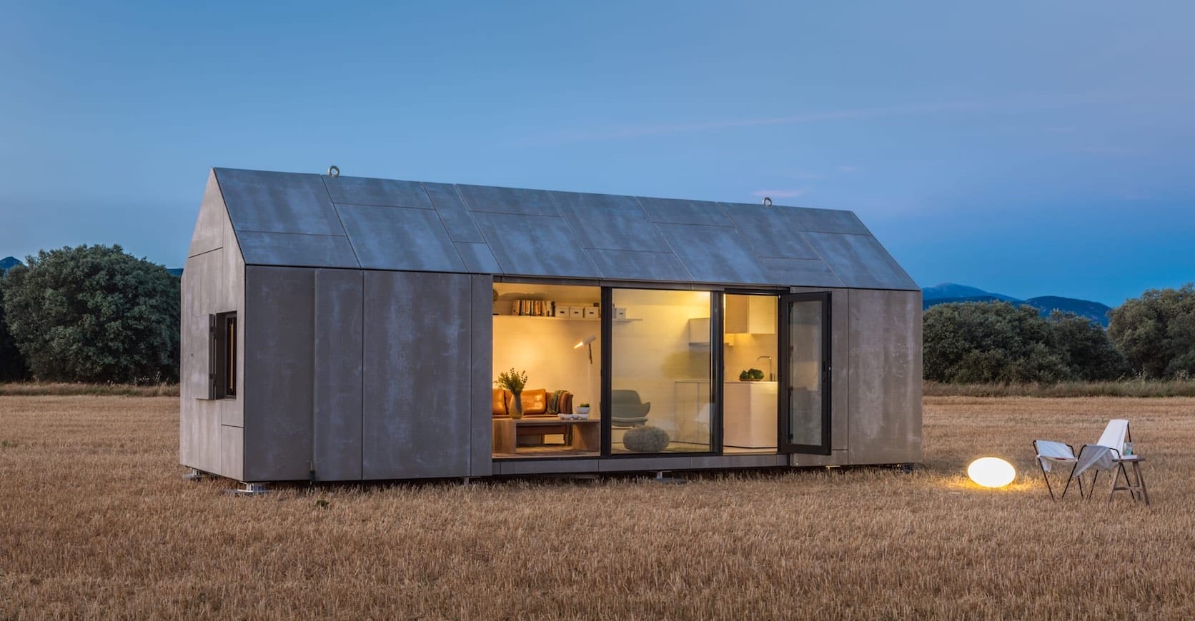 Modernes Modulhaus auf einem Feld, mit erleuchtetem Innenraum und offener Glasfront in der Abenddämmerung