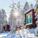 Mehrere Baumhaus-Suiten des Arctic Treehouse Hotels in winterlich verschneiter Waldlandschaft, das Sonnenlicht scheint durch