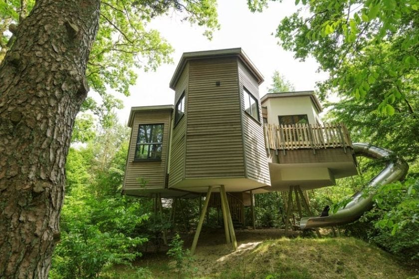 Modernes Baumhaus im Center Parc Bispinger Heide mit Holzfassade, Balkon und Rutsche, umgeben von grünen Blättern