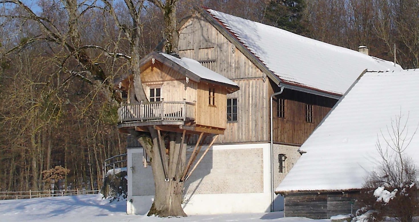 Ein Holz-Baumhaus mit Balkon in einem großen Baum neben einem Gebäude mit schneebedecktem Dach in winterlicher Landschaft