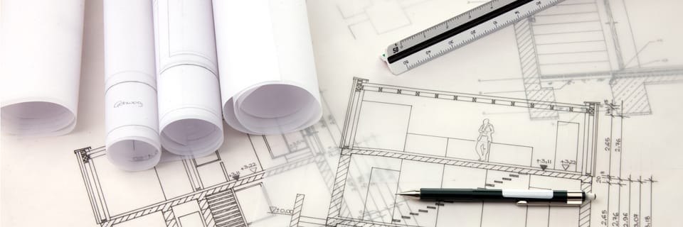 Gerollte Baupläne, daneben ausgebreitete Bauzeichnungen mit eingetragenen Maßen, darüber ein langes Lineal und ein Stift