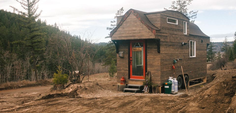 Ein Tiny House aus Holz mit roter Tür, umgeben von Wald und einem schlammigen Weg