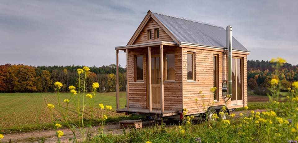 Ein Tiny House aus Holz steht auf einem großen Grundstück mit Wiese und Bäumen, umgeben von gelben Blumen