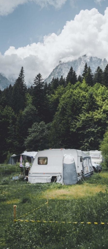 Ein Wohnwagen und ein Zelt auf einem Campingplatz vor einer Bergkulisse mit bewaldeten Wäldern und Gipfeln