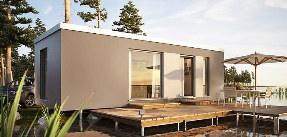 Modernes, quaderförmiges Minihaus am See mit Holzterrasse, Gartentisch und Sonnenschirm, umgeben von Bäumen und einem Auto