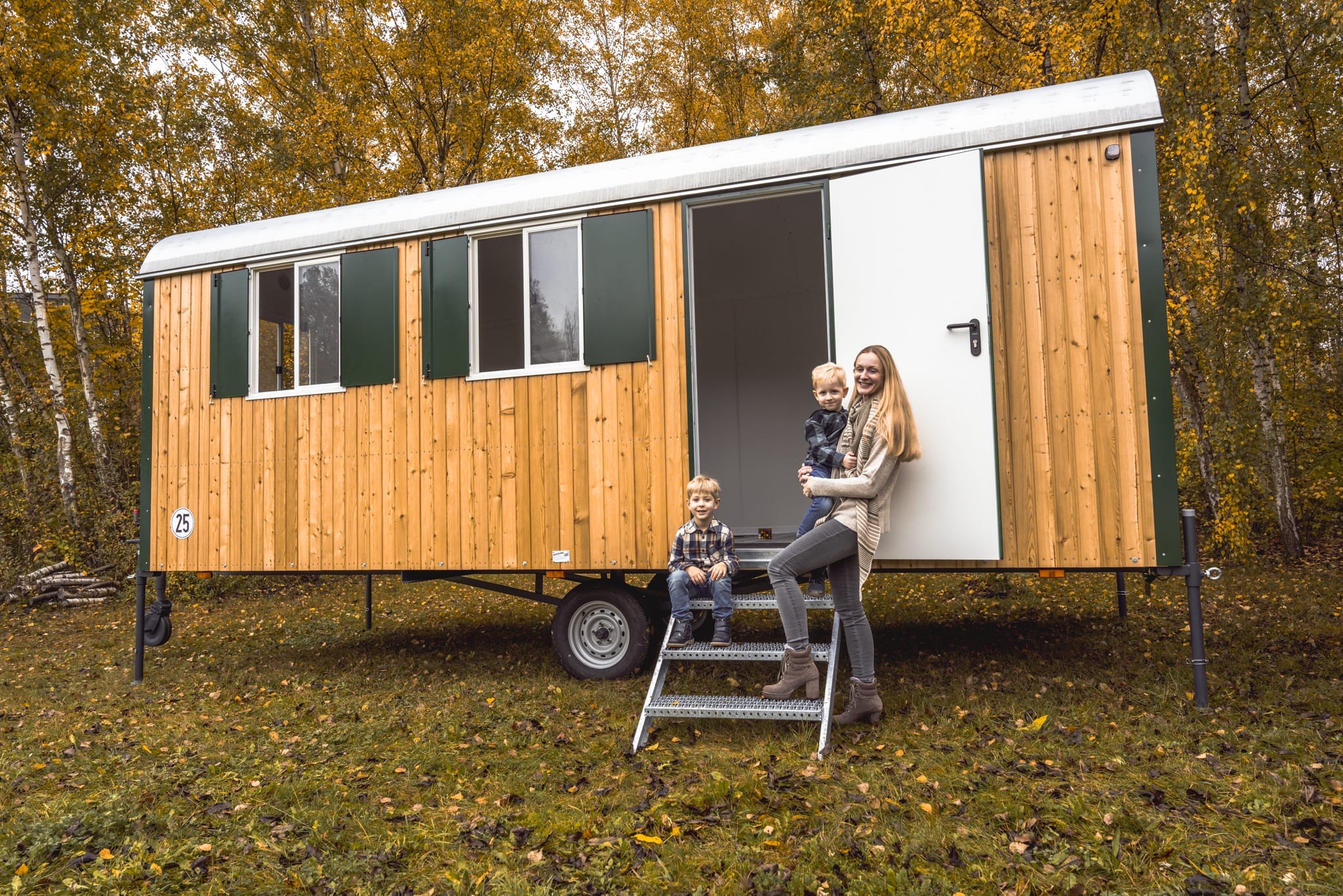 Eine lachende Frau mit zwei kleinen Jungen posiert vor einem dunkelgrünen Holzwohnwagen im Herbstlaub