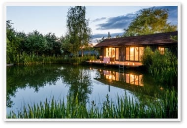 Ein Holzhaus am Teich mit beleuchteten Fenstern und einer Terrasse, umgeben von grünen Pflanzen bei Dämmerung