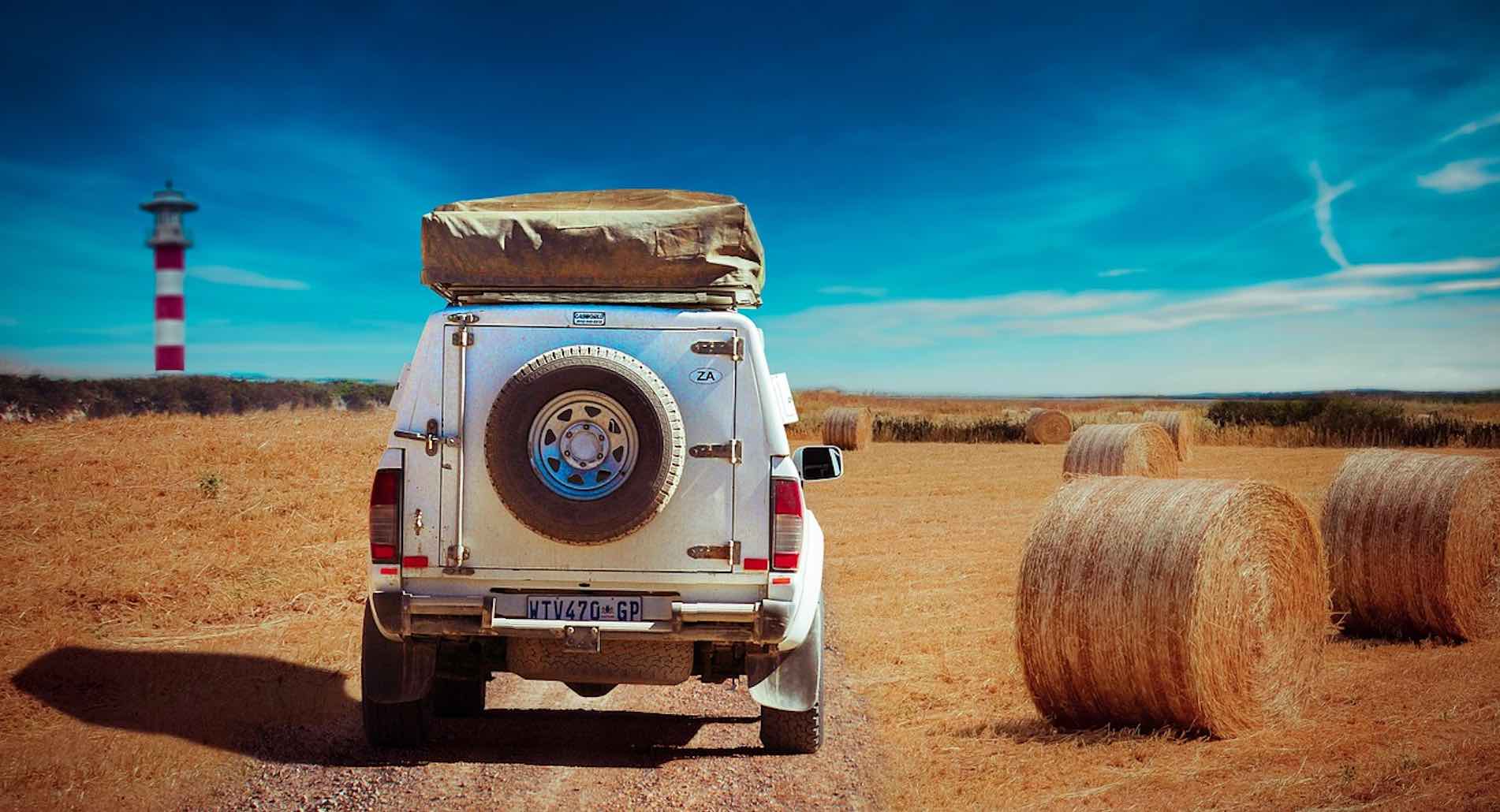 Geländewagen mit Dachzelt auf einem Feldweg umgeben von Strohballen und einem Leuchtturm am Horizont
