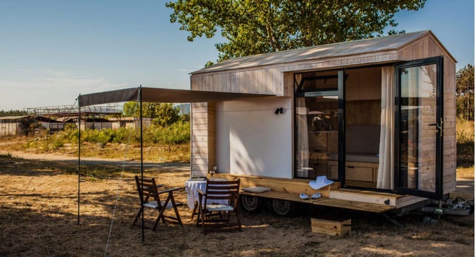 Ein Tiny House aus Holz mit kleiner Terrasse, davor unter einem Sonnensegel zwei Stühle und ein kleiner Tisch