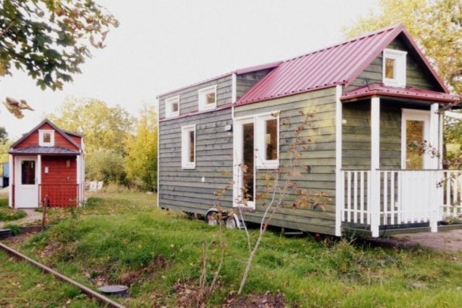 Ein hellgrünes Tiny House mit rotem Dach und Veranda steht auf einer Wiese, dahinter ein kleineres rotes Tiny House