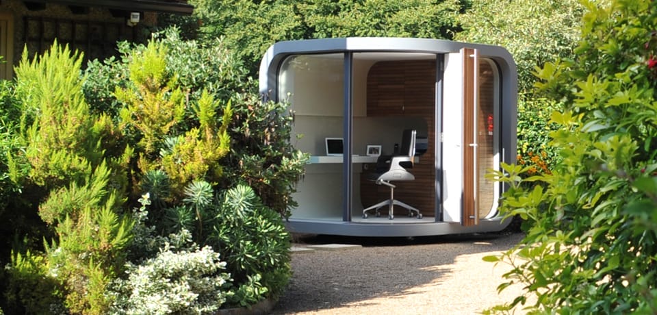 Ein Garten-Studio mit runden Ecken und Glasfront steht inmitten von grünen Pflanzen, davor ein Schotterweg