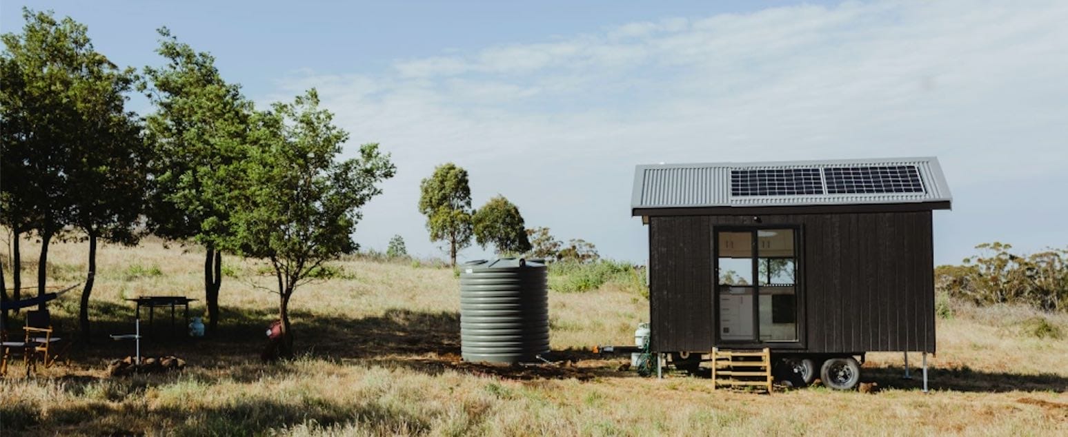 Tiny House mit Solarpaneelen auf einem Grundstück mit Wiese, Bäumen und Wassertank