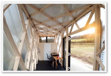 Innenansicht kleines Tiny House aus Holz mit Blick auf Felder und strahlende Sonne am Horizont stehende Sonne