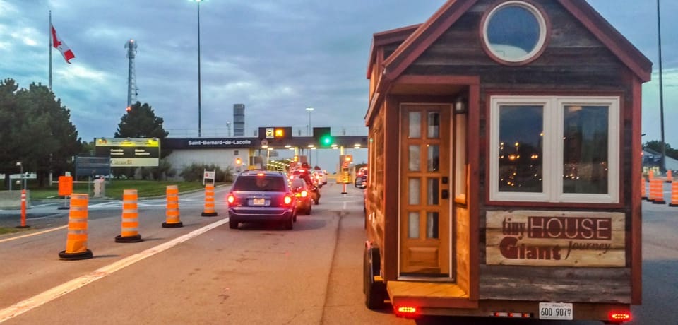 Ein Tiny House auf Rädern vor einer Mautstelle mit parkenden Autos und Hinweisschildern von Saint-Bernard-de-Lacolle
