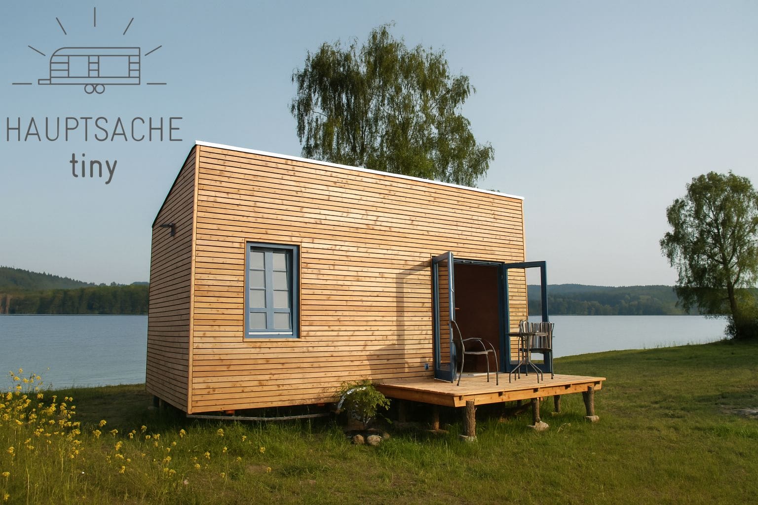 Ein Tiny House am Seeufer vor bewaldeter Hügelkette und blauem Himmel. Oben links ein Logo mit Schriftzug "HAUPTSACHE tiny"