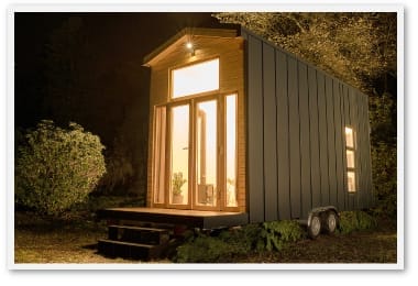 Ein modernes Minihaus mit großen Fenstern und Holzverkleidung bei Nacht, das innen hell erleuchtet ist