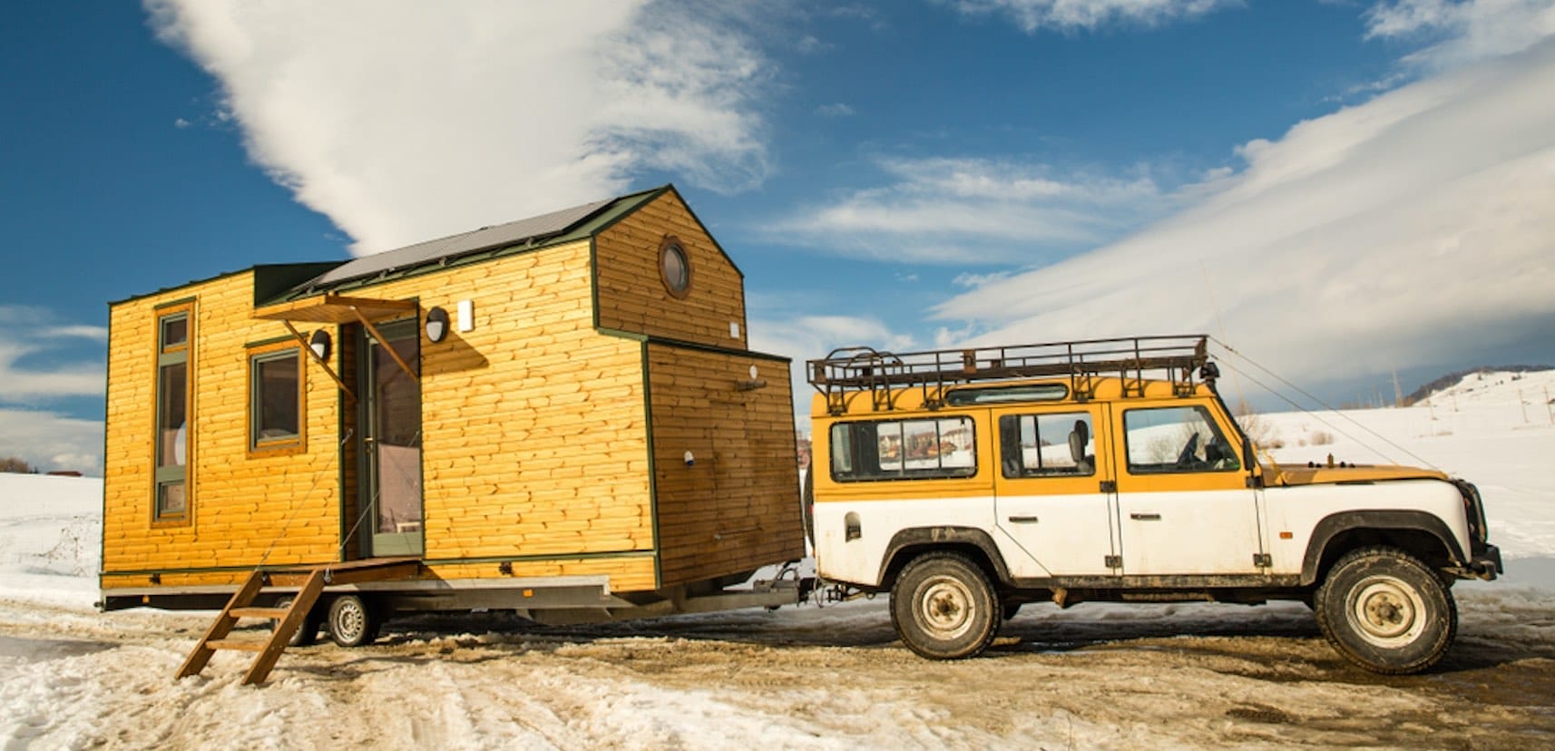Ein gelb-weißer Geländewagen zieht ein kleines Holzhaus mit Solarpaneelen über eine schneebedeckte Landschaft