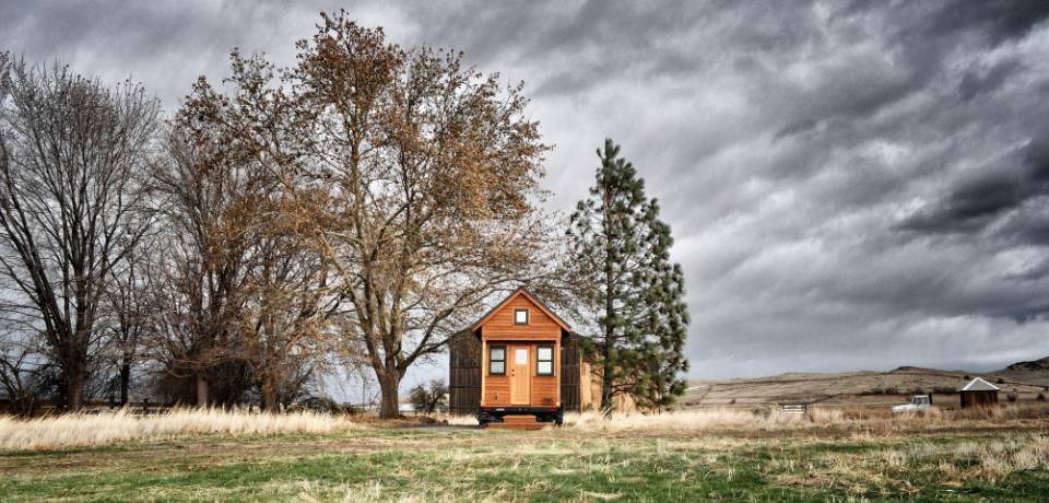Holz-Tiny-House auf Anhänger zwischen Bäumen auf einer Wiese unter grauem Himmel