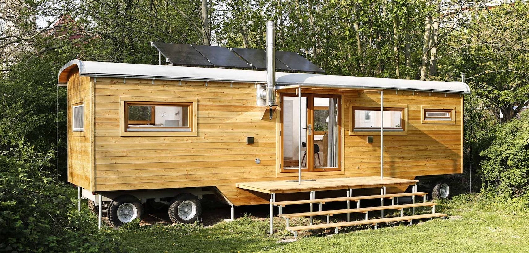 Holzverkleideter Tiny House on Wheels mit Terrasse, Solarkollektoren und Schornstein, umgeben von Bäumen und Grünfläche von d